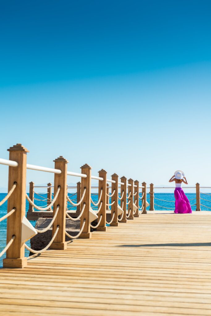 A wooden pier in Larnaca, Cyprus, extends into the clear blue sea under a vast sky. Decorative railings line the pathway, and a woman in a bright pink dress with a white hat gazes out at the expansive seaview, embodying the serene coastal atmosphere.