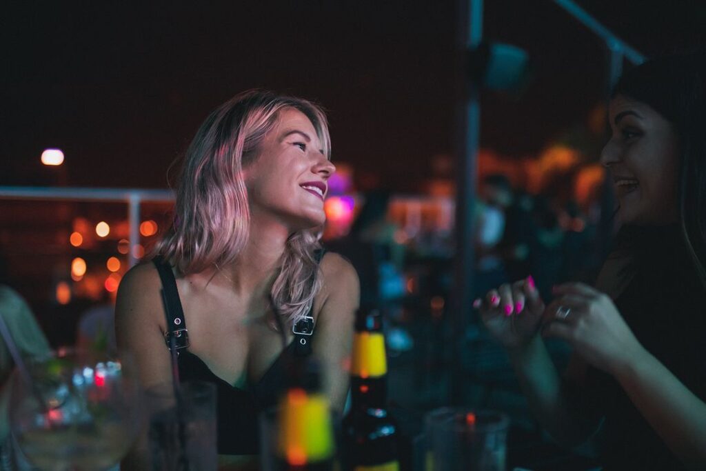 Two women enjoying an evening at Memories rooftop bar in Larnaca; one with pink-tinted hair looking up and the other talking animatedly, surrounded by dim lighting and bar essentials