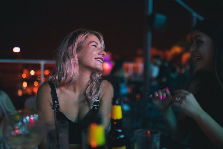 Two women enjoying an evening at Memories rooftop bar in Larnaca; one with pink-tinted hair looking up and the other talking animatedly, surrounded by dim lighting and bar essentials